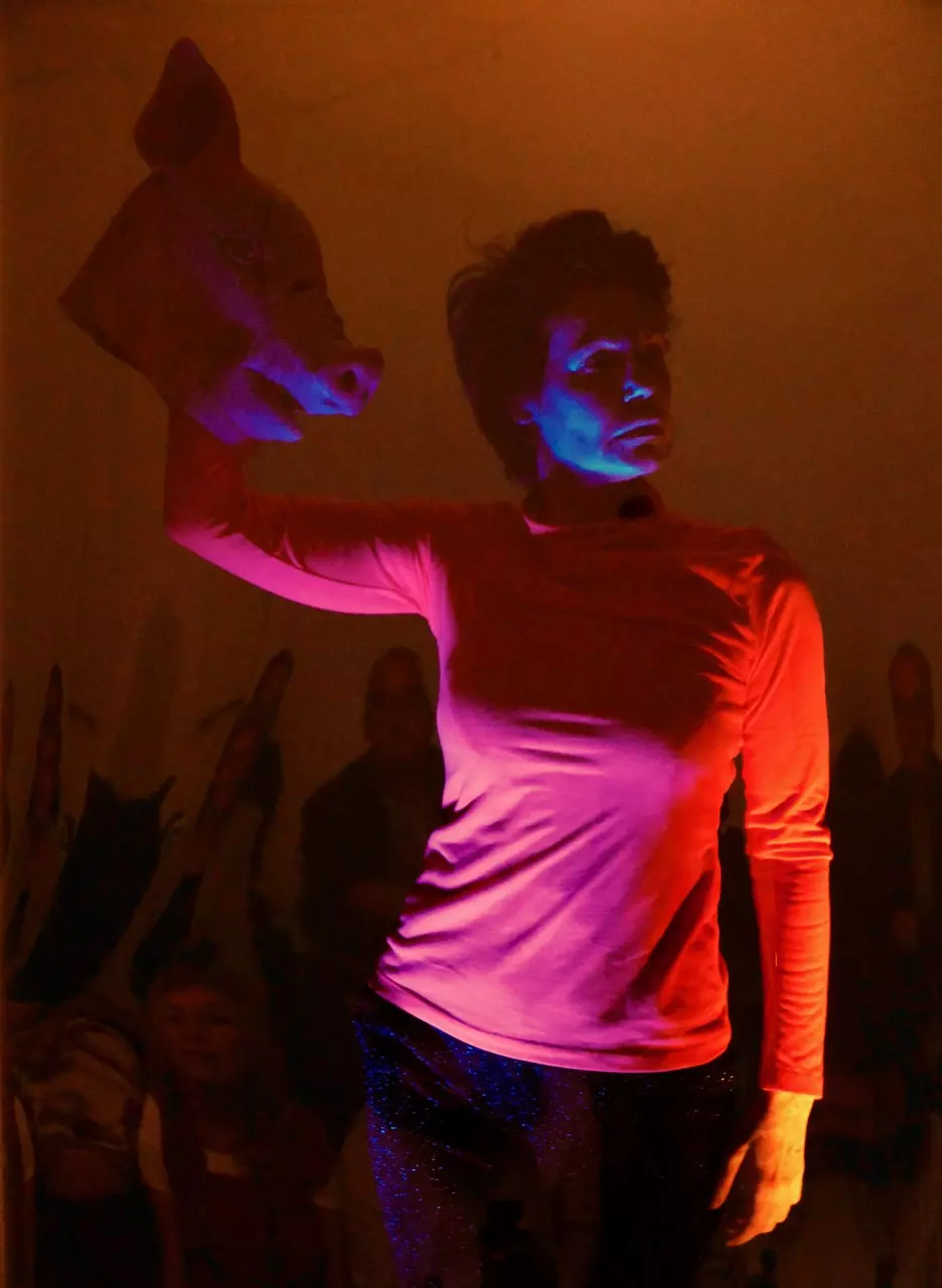 Gerda Brodmann-Raudonikis, eine Frau mit schwarzen kurzen Haaren, schaut zur Seite. Ihr rechter Arm ist angewinkelt und trägt eine Schweinemaske