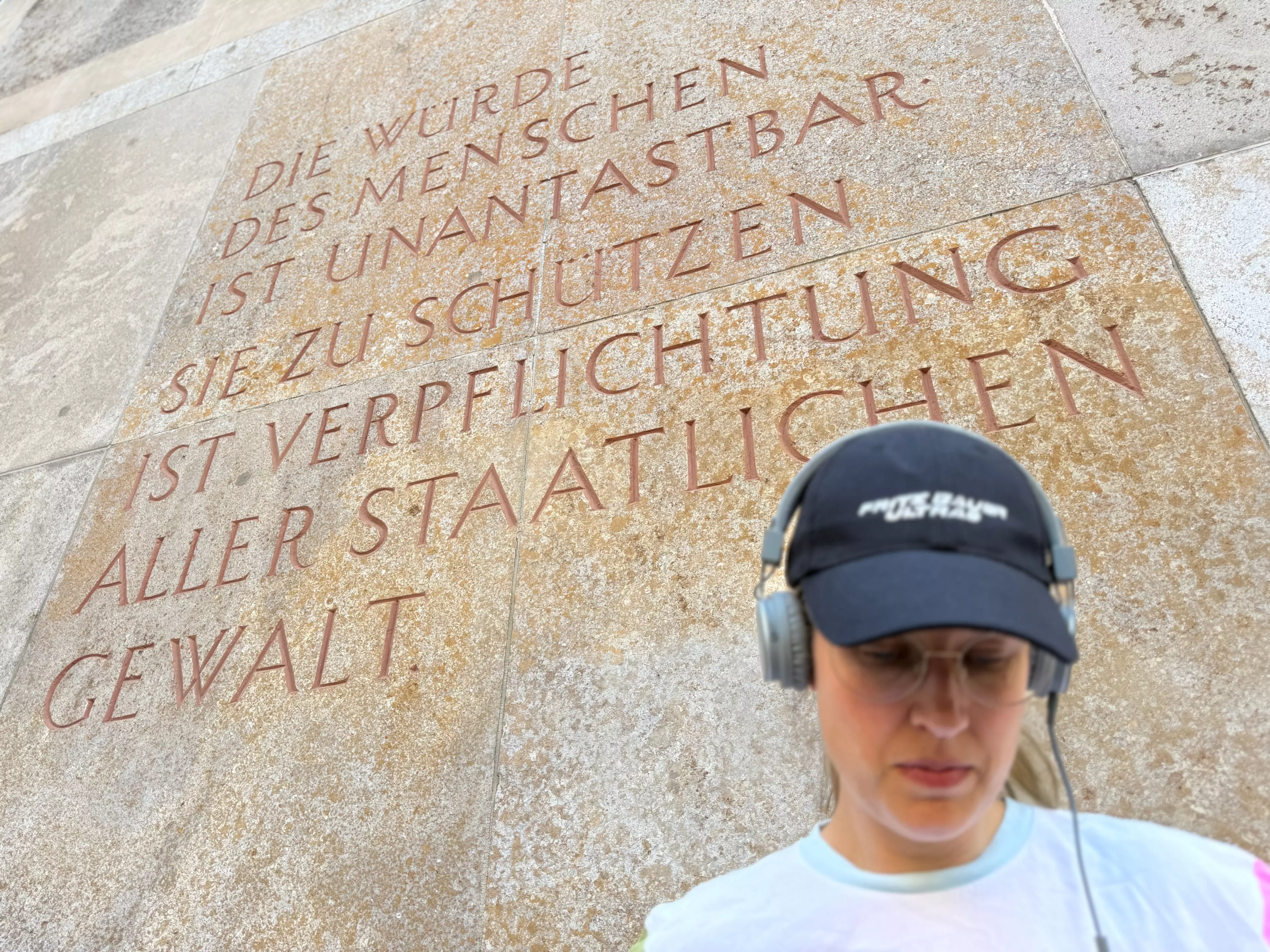Eine Person mit leicht gesenktem Kopf - nachdenklich. Sie trägt ein Cap mit der Aufschrift "Fritz Bauer Ultras" und Kopfhörer. Im Hintergrund an der Wand, der 1. Artikel des Grundgesetzes.