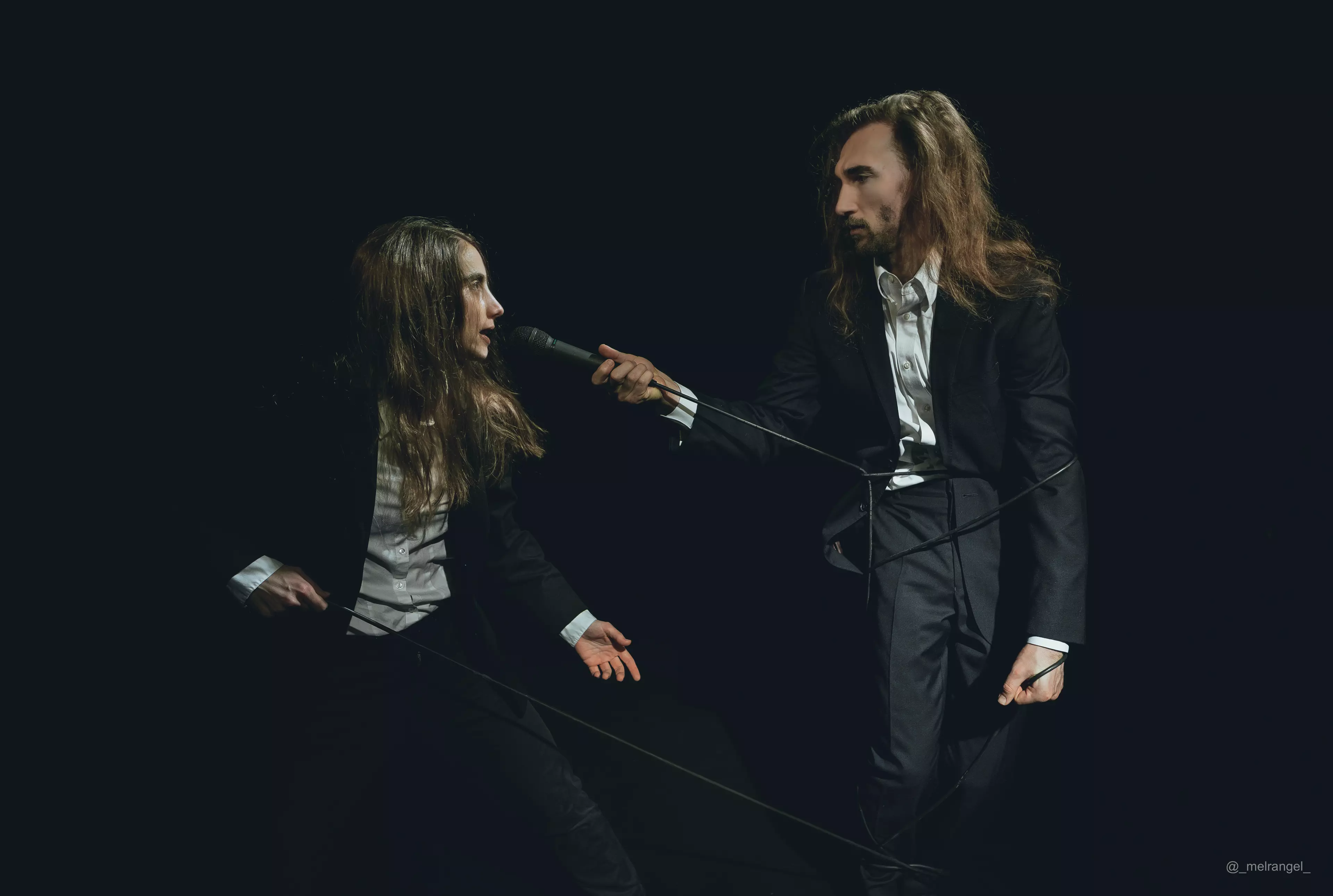 Eine junge Frau (Alina Jaggi) und ein junger Mann (Joshua Haines) stehen sich gegenüber. Sie tragen beide einen schwarzen Anzug und ein weißes Hemd. Alina zieht an einem Kabel, das um Joshua gewickelt ist. am anderen Ende des Kabels ist ein Mikrofon, dass Joshua Alina vor den Mund hält.