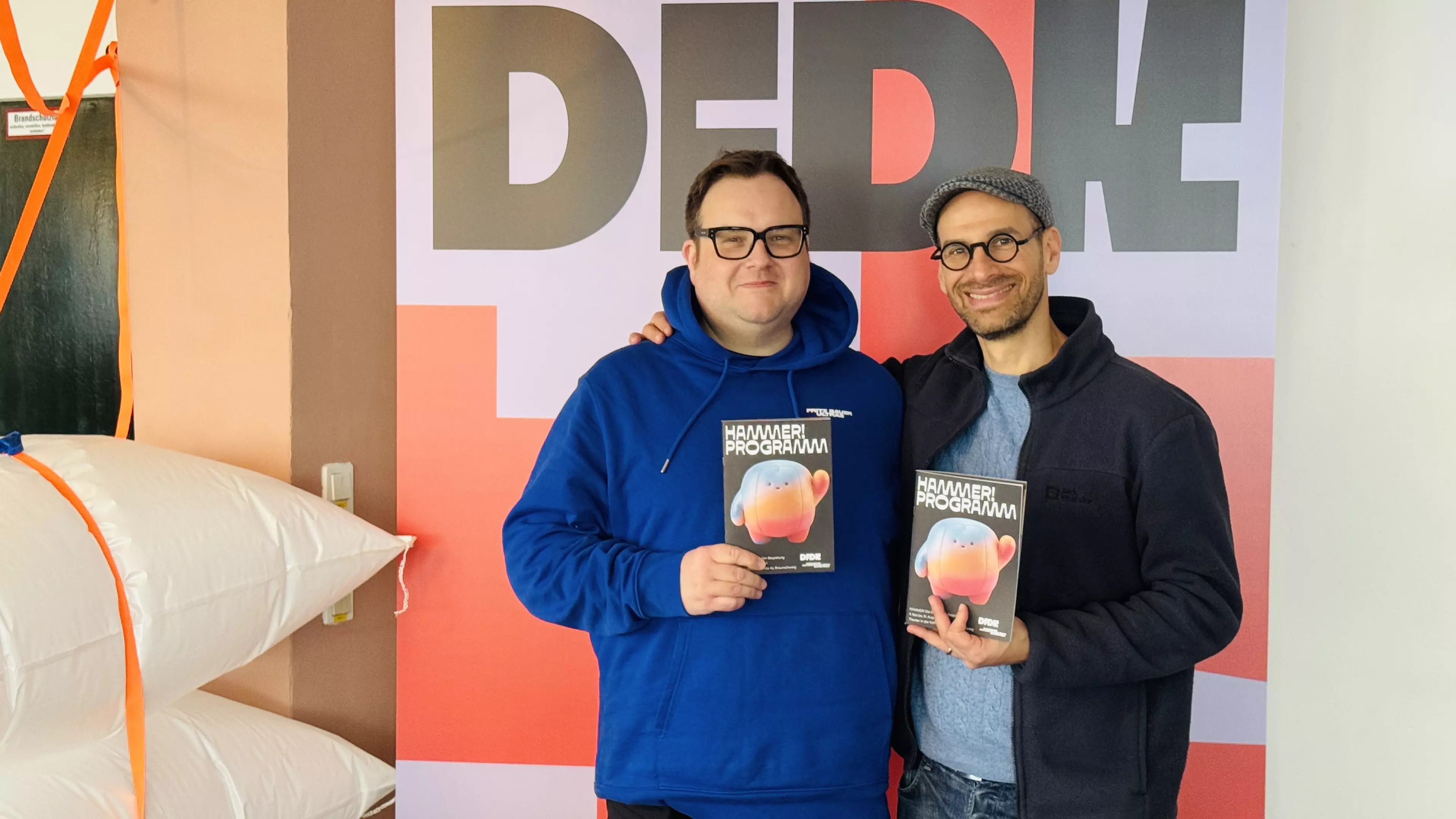 Zwei Personen (Christian Weiß & Fabian Cohn), die freundlich in die Kamera blicken und jeweils ein Programmheft der Spielreihe in die Kamera halten.