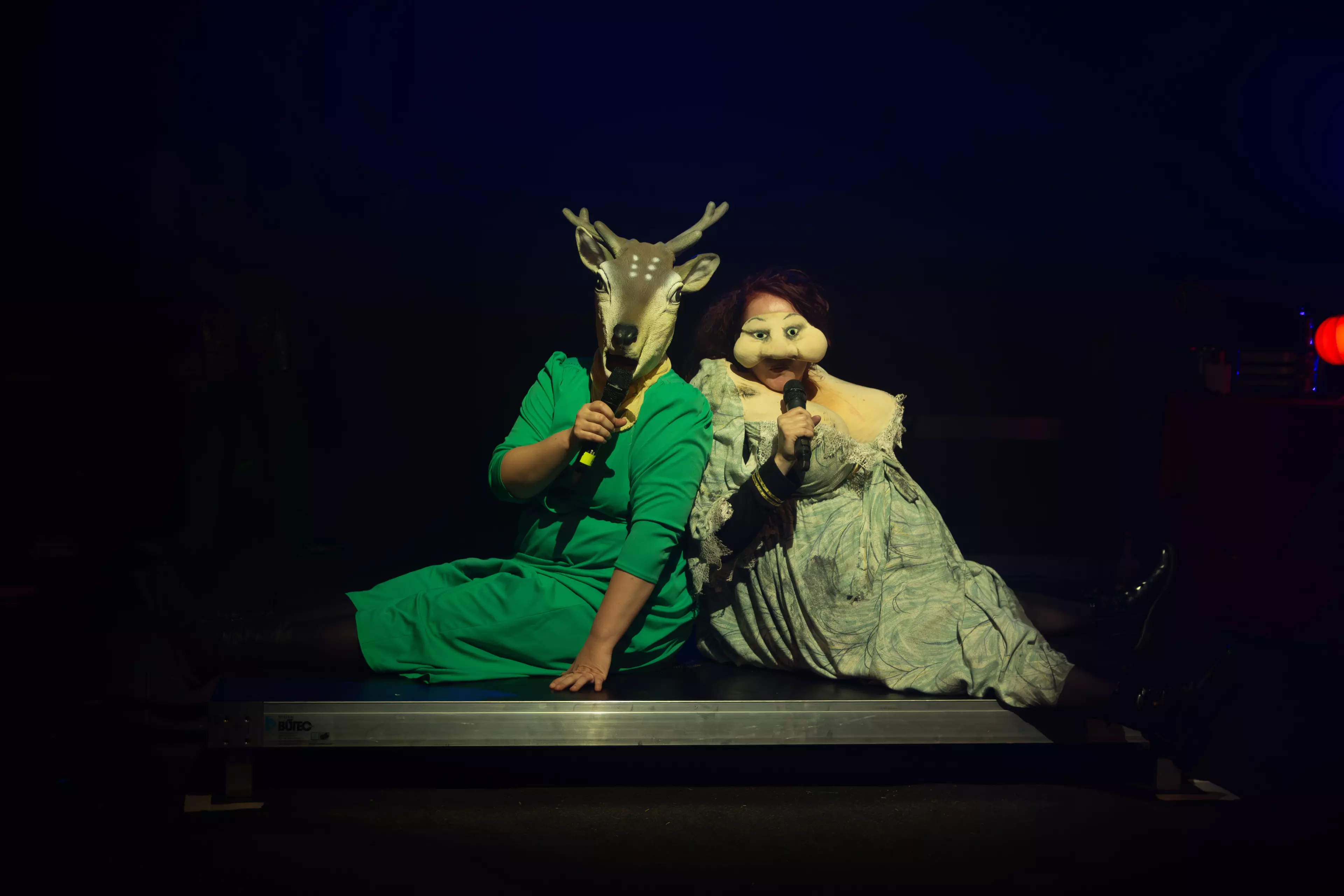 Eine Frau im grünen Kleid mit einer Reh Maske sitzt neben einer Frau im grauen Kleid mit einer Halbmaske die ihr dicke Wangen macht. Beide halten ein Mikro in der Hand.