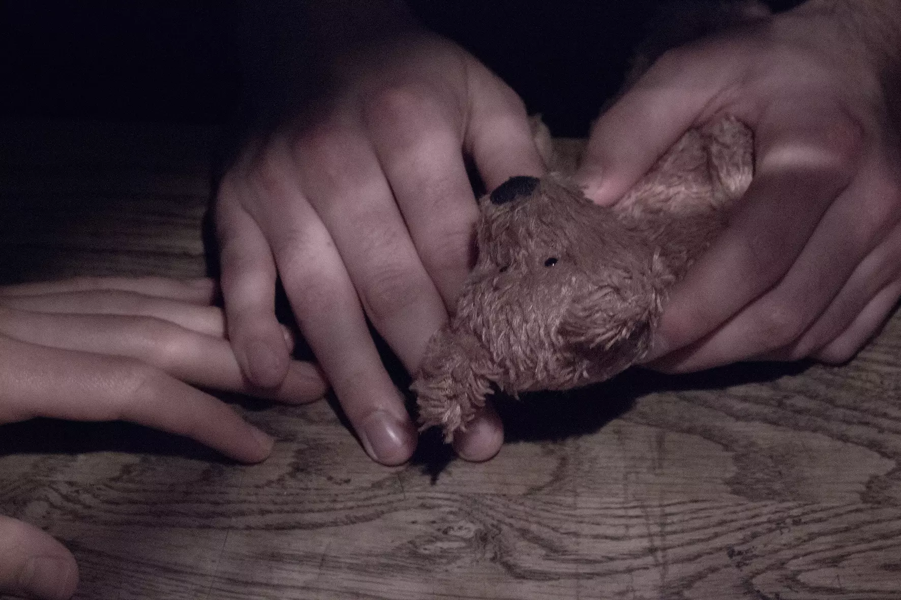 Zwei Hände, die auf einem Tisch liegen und einen kleinen Teddybär halten. Einige Finger berühren eine dritte Hand.