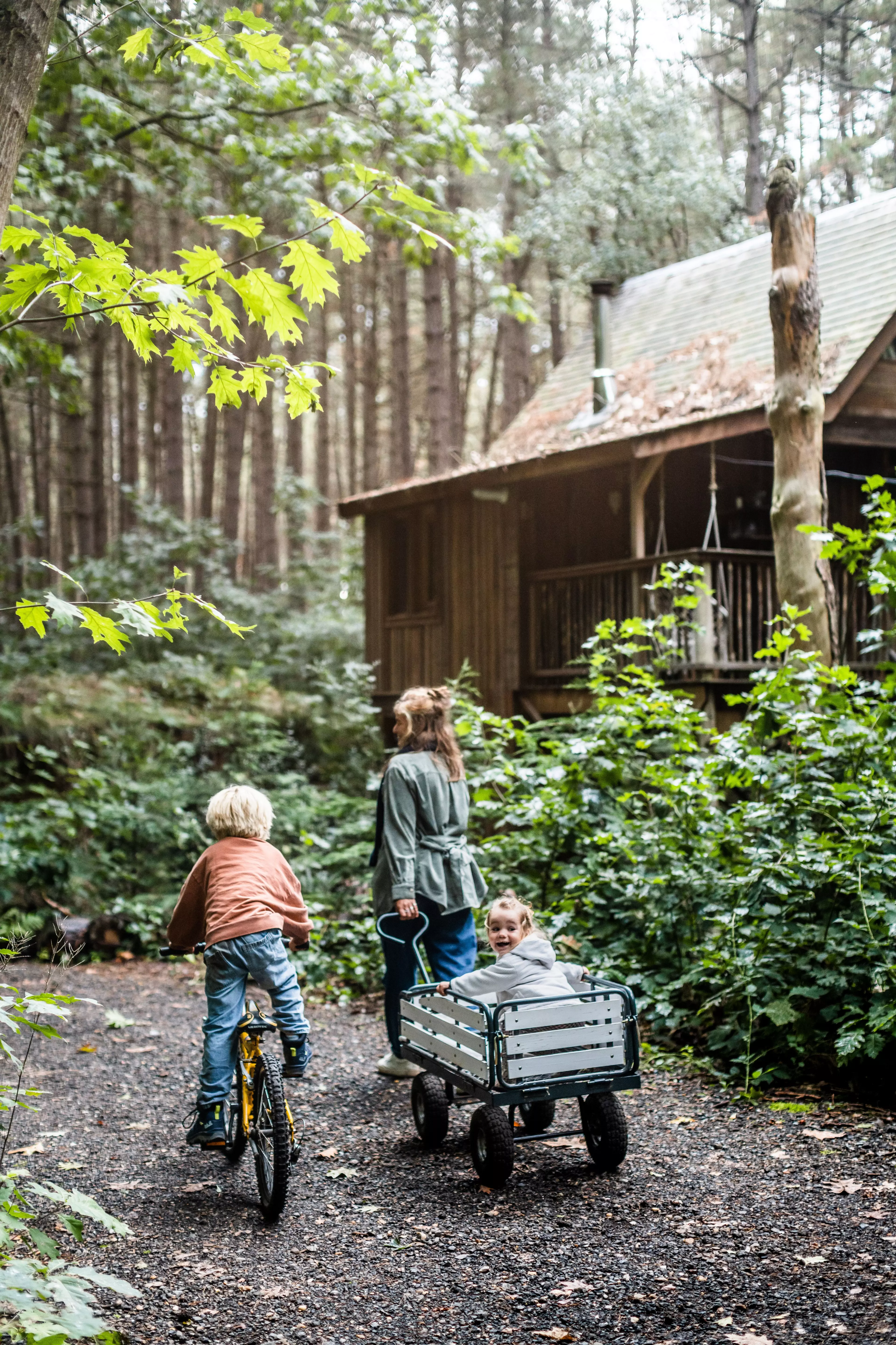 Ein Kind faehrt Fahrrad, ein zweites wird in einem hoelzernen Bollerwagen gezogen – die Treelodge in Belgien liegt mitten in einem Erlebniswald.