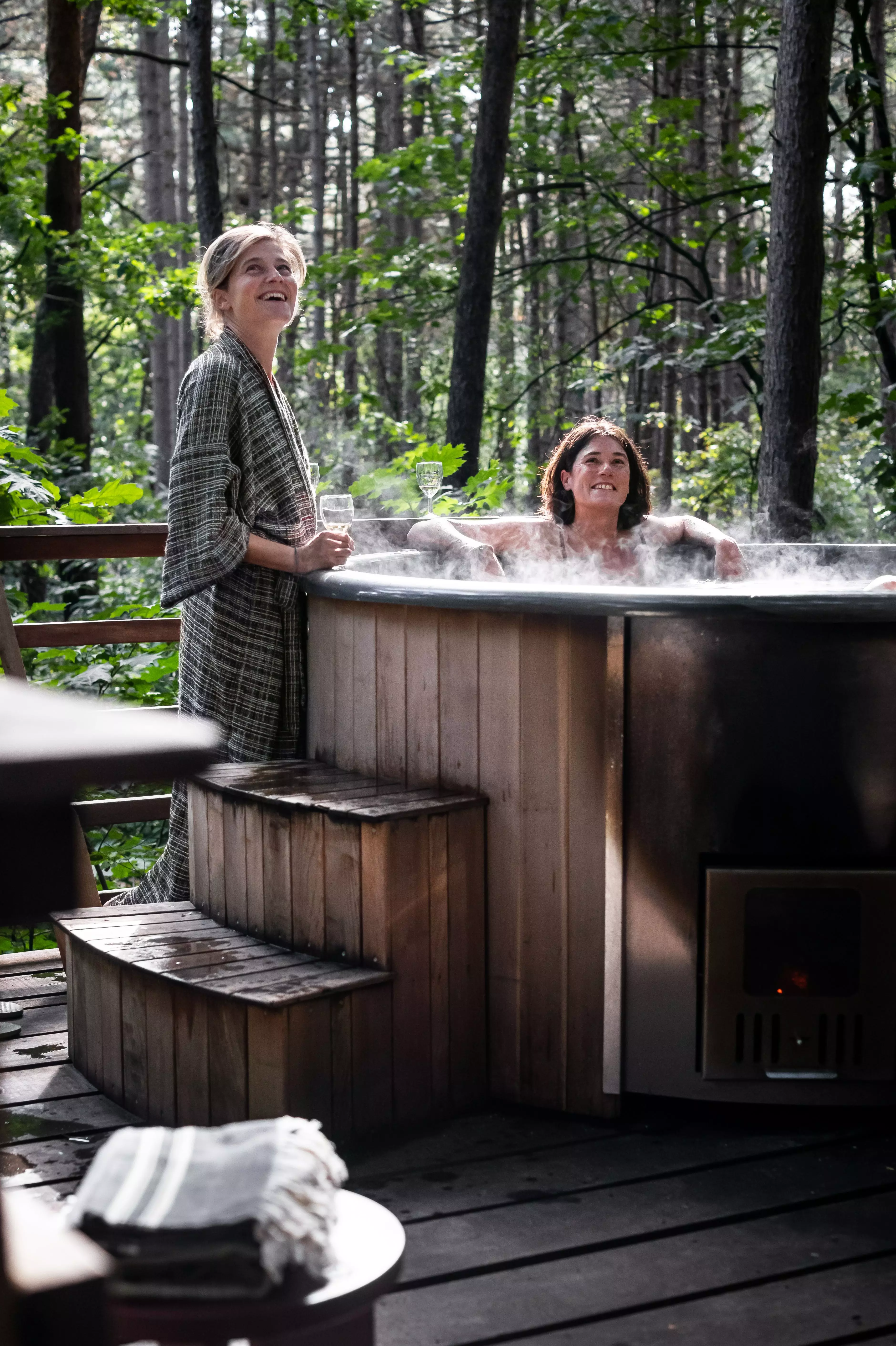 Eine Frau sitze in einem hoelzernen Aussenpool der Treelodge in Belgien, eine steht im Bademantel daneben.