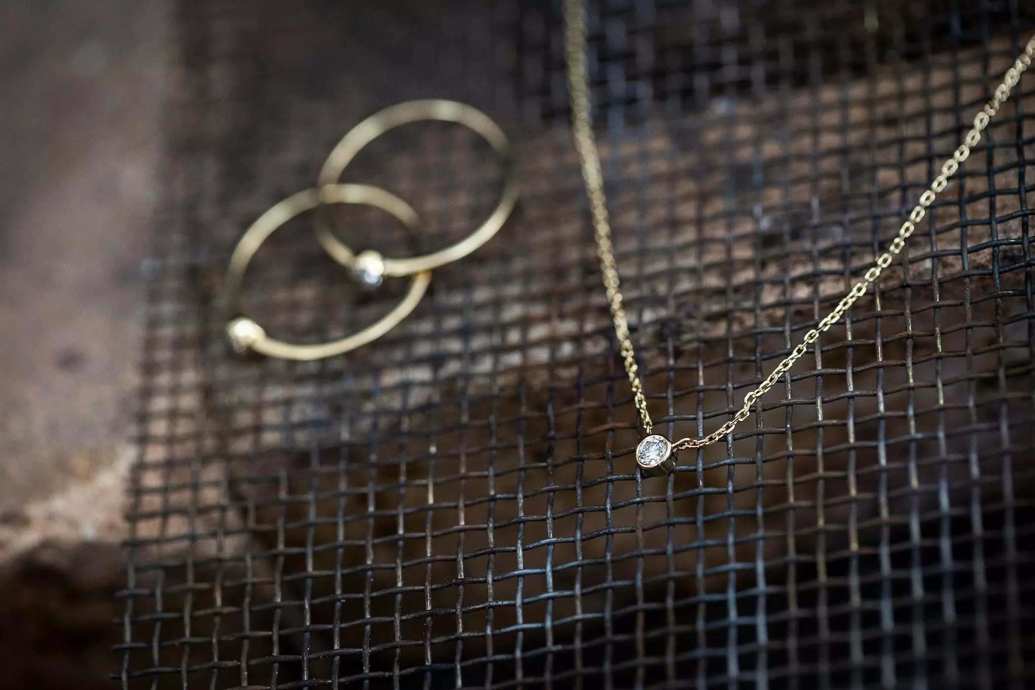 Im Vordergrund eine zarte Goldkette mit einem runden Diamant, im Hintergrund zwei zarte Goldringe mit jeweils einem Solitaire-Stein, auf einem Metallgitter liegend – femtastics