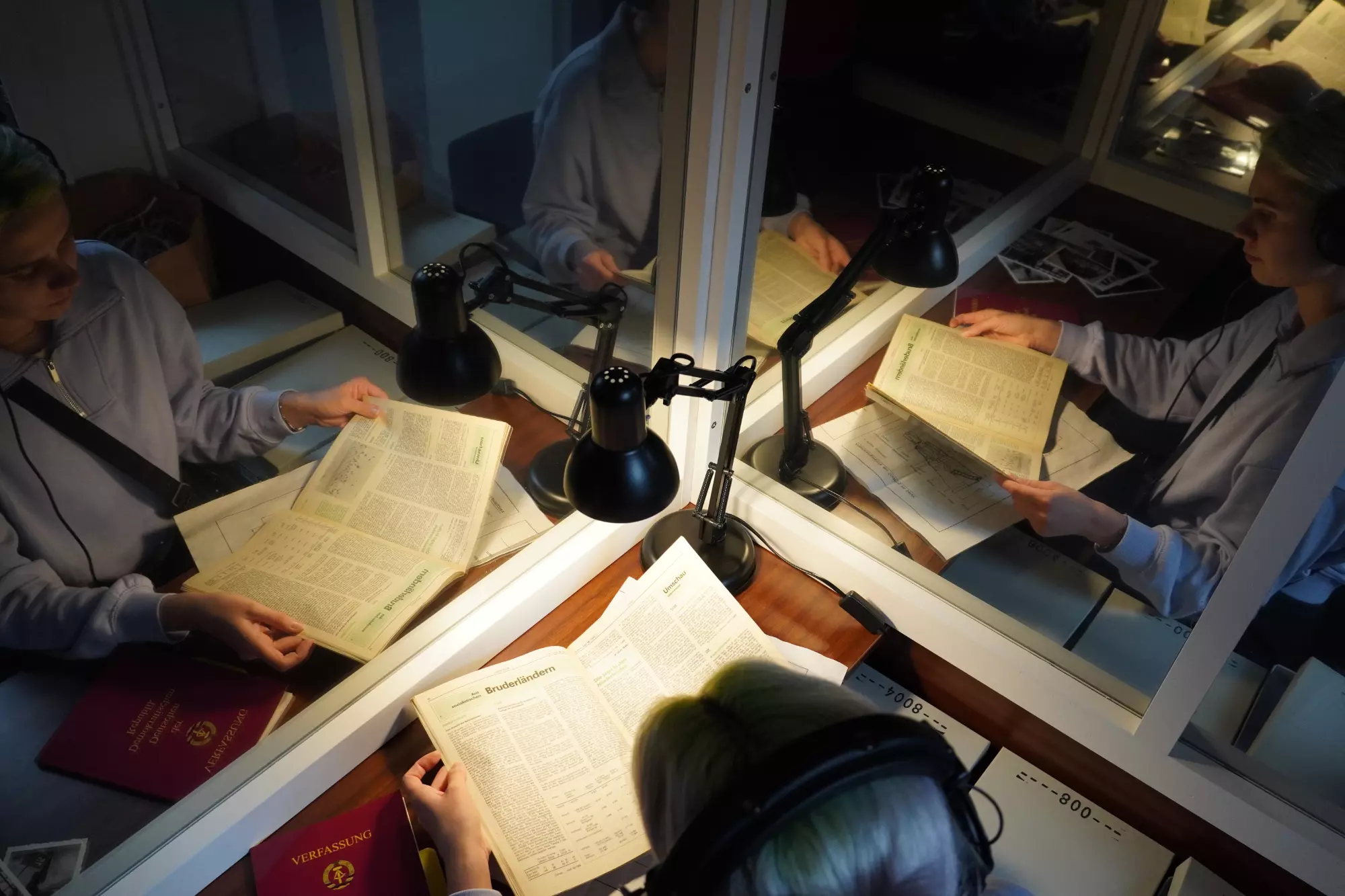Ein Tisch in einer Glaskabine. Die Schreibtischlampe leuchtet und der Hinterkopf einer Person ist sichtbar, die Unterlagen vor sich auf dem Tisch hat und sie durchschaut.