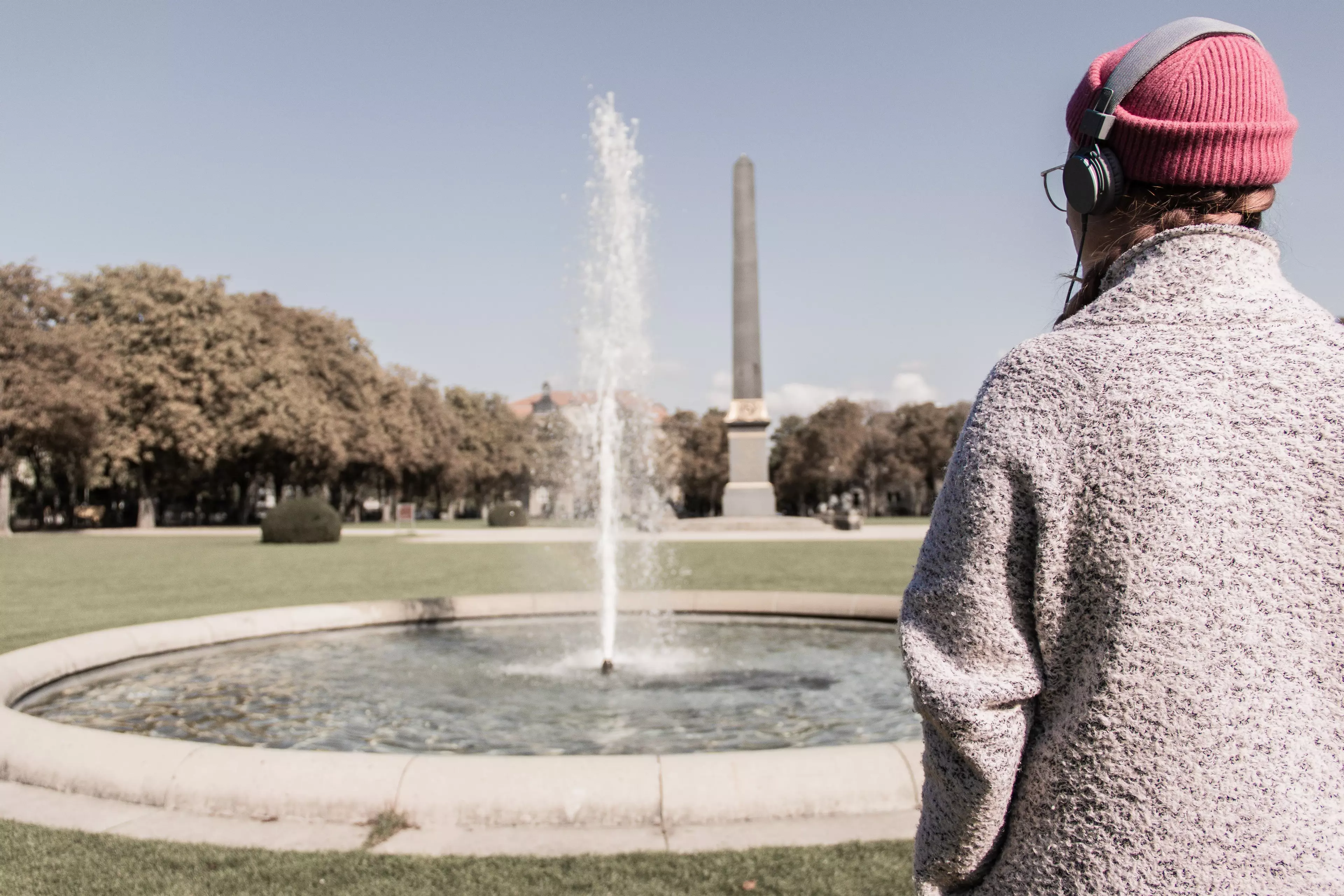 Im rechten Bildvordergrund eine weiblich gelesene Person in Rückenansicht. Sie trägt einen Mantel, eine rosafarbene Mütze und Kopfhörer. Sie blickt in die Bildmitte: eine Wasserfontäne und ein Obelisk im Hintergrund.