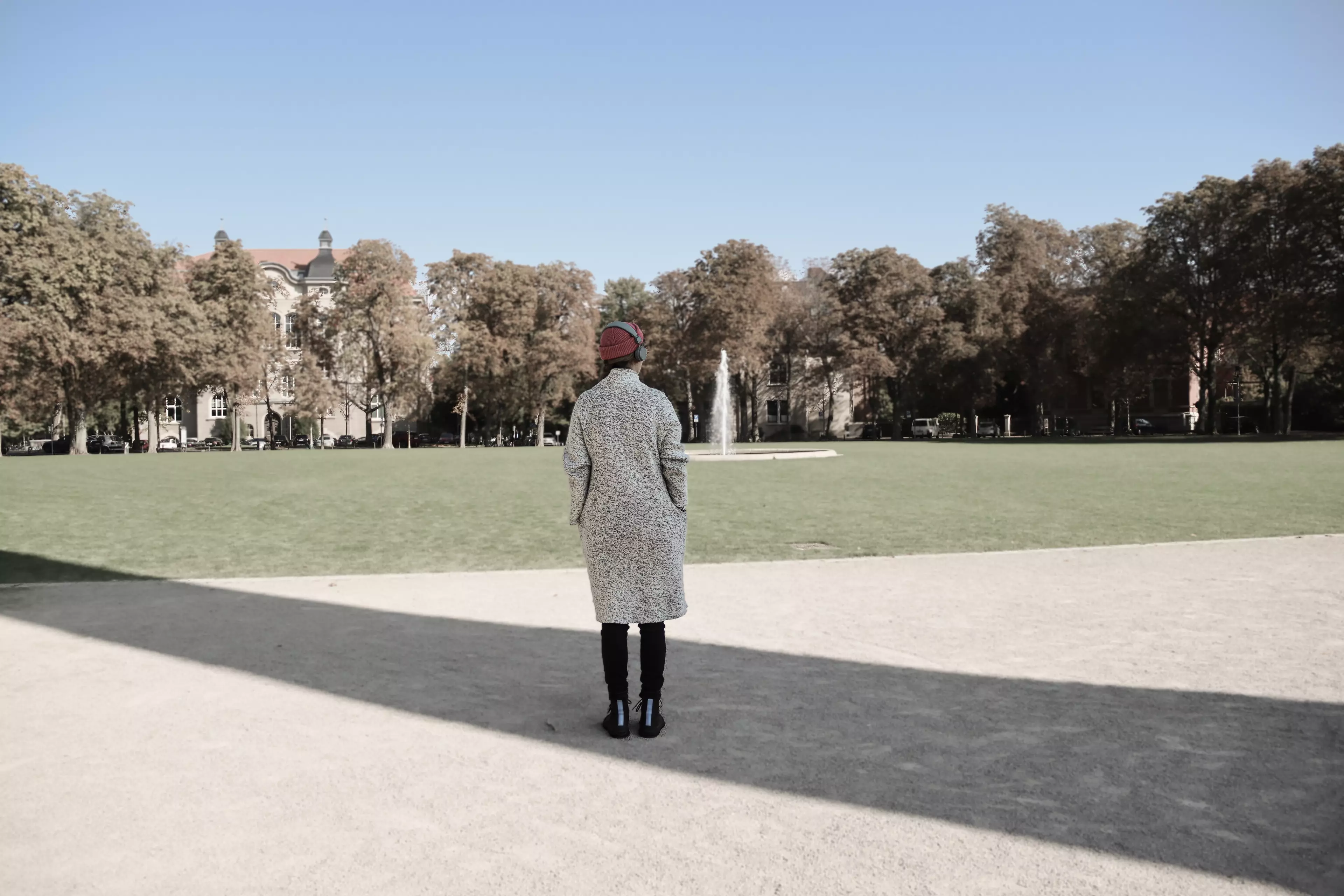 Ein nichtbares Objekt wirft einen langen Schatten ins Bild. Eine weiblich gelesene Person steht mit dem Rücken zu uns in diesem Schatten.